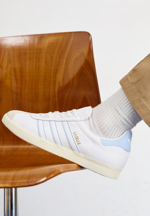 Biały sneaker Adidas Gazelle z jasnoniebieskimi paskami i piętą, noszony z białymi skarpetami i beżowymi spodniami, odpoczywający na drewnianym krześle.