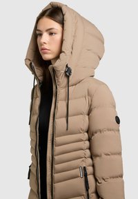 Tan pufferjakke med en stor hætte, lodret quiltet design, lynlåslommer og justerbare snoretræk på hætten.
