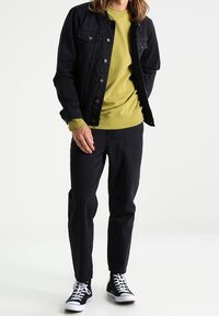 Černá džínová bunda, zelený svetr, černé kalhoty a černé tenisky. Bunda má knoflíkové kapsy a outfit má ležérní design.