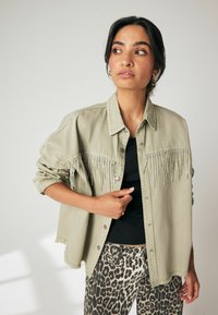 Šviesiai khaki džinsinis švarkas su frangėmis ir mažais karoliukais, turintis apykaklę ir suplyšusį hemą, derinamas su leopardų raštuotomis kelnių.