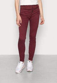 Jean skinny bordeaux avec boutons sur le devant, en denim doux, à taille mi-haute, associés à des baskets blanches et roses.