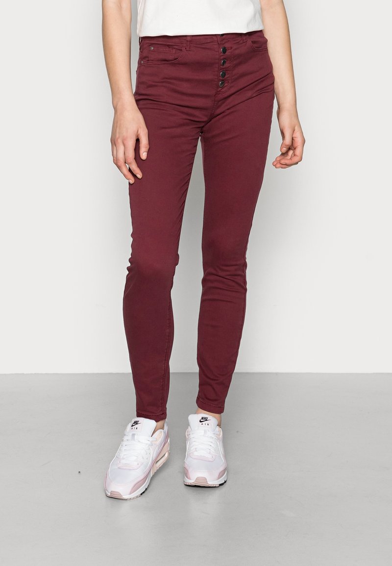 Jean skinny bordeaux avec boutons sur le devant, en denim doux, à taille mi-haute, associés à des baskets blanches et roses.