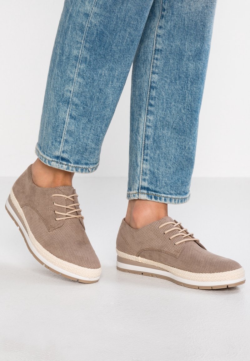 Chaussures décontractées beiges à lacets avec côtés texturés, portées avec un jean en denim bleu clair, présentées sur un fond blanc uni.