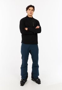 Zwarte, met rits, langemouwen top; blauwe skibroek; zwarte skibotten. Eenvoudig ontwerp met gestructureerde stof en aansluitende silhouet.