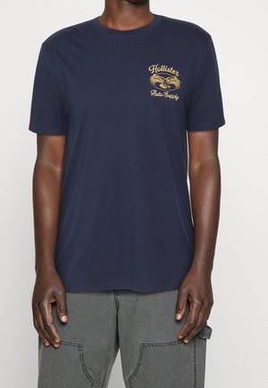 Person iført en mørkeblå Hollister T-shirt med et gult ørnelogo og teksten "Auto Supply" på brystet, kombineret med grå bukser.