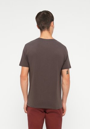 Mand vendt væk iført en enkel mørkegrå kortærmet t-shirt og røde bukser med en lille pil-tatovering på venstre arm.