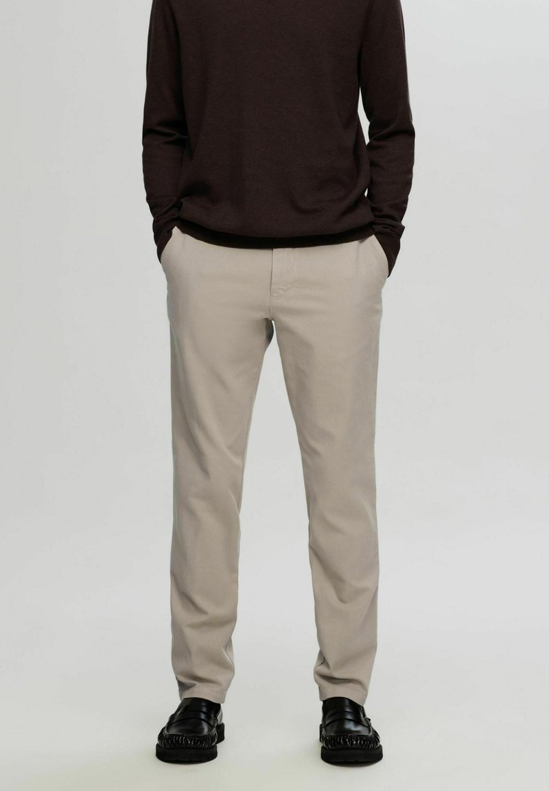 Chino beige con vestibilità dritta, realizzati in un morbido misto cotone. Abbinati a un maglione scuro e mocassini neri, senza motivi visibili.