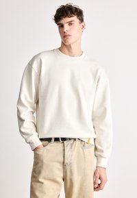 Κρεμ ελαιοκόκκινη μπλούζα με μακριά μανίκια και στρογγυλή λαιμόκοψη. Φορεμένη με ανοιχτόχρωμο μπλε τζιν και μαύρη ζώνη. Απλός σχεδιασμός, μαλακή υφή.