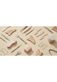 Vari strumenti per la cura della pelle in oro rosa e rosa, tra cui pietre gua sha, pinzette, spazzole e dispositivi per la pulizia del viso, disposti su una superficie beige.
