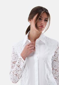 Weißes Hemd mit Spitzenakzenten an den Ärmeln und Seiten, klassischem Kragen, durchgehender Knopfleiste, glatter Textur und aufwendigem Blumenmuster.