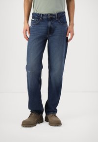 Blue denim jeans with a straight-leg design, faded texture, distressed knee detail, and five pockets. Worn with brown lace-up boots.