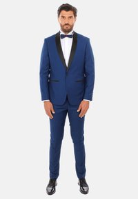 Tuxedo blu con revers in satin nero, forma aderente, pantaloni a gamba dritta e scarpe nere lucide. Camicia bianca con papillon.
