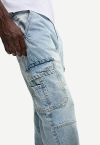 Svijetloplave traper cargo hlače s više džepova, vidljivim šavovima i opuštenim krojem. Tkanina izgleda mekano s blago izblijedjelim završetkom.