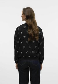 Svart sweatshirt med vita rosett-mönster, ribbade mudd och nederkant, avslappnad passform, bärs med mörka jeans, sedd bakifrån.