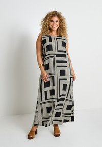 Robe maxi avec un motif géométrique en noir et crème, design sans manches, tissu léger et coupe décontractée. Portée avec des sandales à talons bruns.