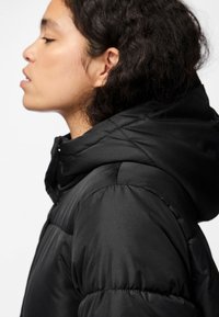 Casaco puffer preto com capuz, apresentando um design acolchoado, tecido texturizado e detalhe de costura proeminente nos ombros e mangas.