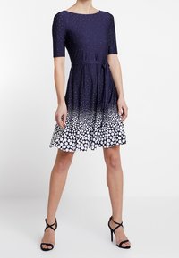 Kratší rukáv tmavě modré šaty s kulatým výstřihem, zdobené bílým puntíkovaným vzorem, který přechází od malých puntíků k velkým, a s vázaným pasem.