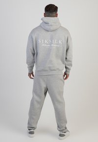 Sivá mikina s kapucňou a logom "SIKSILK" na chrbte, spárovaná so zodpovedajúcimi sivými teplákmi. Vyrobené z mäkkej, textúrovanej zmesi bavlny.