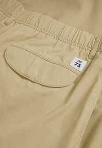 Shorts kaki avec une taille élastique et une seule poche arrière arrondie. Présente une étiquette en tissu avec l'inscription "M 73" cousue dessus.