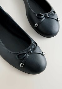 Schwarze Ballerinas aus strukturiertem Material mit runder Zehenpartie, einer Schleifenakzent mit Metalldetail und einem glatten Innenfutter.