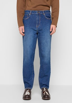 Uomo che indossa jeans blu con gamba dritta, maglia marrone a maniche lunghe e scarpe nere con dettagli bianchi e marroni a fantasia.