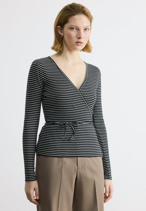 Γυναίκα που φοράει ασπρόμαυρο ριγέ πουλόβερ με ζώνη στη μέση και μπεζ ψηλόμεσο παντελόνι, ποζάροντας μπροστά σε απλό ανοιχτό φόντο.