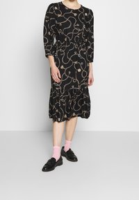 Svart klänning med ett mönster av kedjor och tofsar, trekvartslånga ärmar, samlad midja och en volangnederkant. I kombination med svarta skor och rosa strumpor.