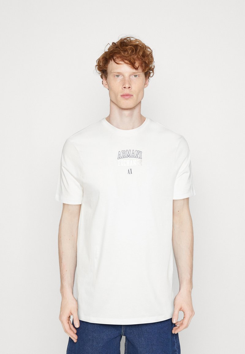 Camiseta de algodón blanca, de manga corta, cuello redondo, con el logo "Armani Exchange" en la parte delantera, combinada con pantalones de denim azules. Textura suave, ajuste relajado.