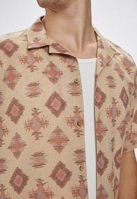 Beżowa koszula z krótkim rękawem z wzorem diamentowym i abstrakcyjnym w odcieniach rdzy i szarości. Posiada naturalne drewniane guziki i luźny kołnierz.