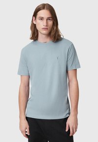 Jasnoniebieska bawełniana koszulka z krótkim rękawem, okrągłym dekoltem i małym logo wyhaftowanym na piersi. Noszona z czarnymi spodniami.