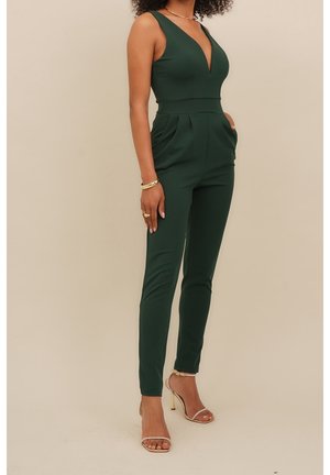 Green fitted jumpsuit with deep V-neck, sleeveless design, side pockets, and tapered legs, paired with strappy high-heeled sandals.