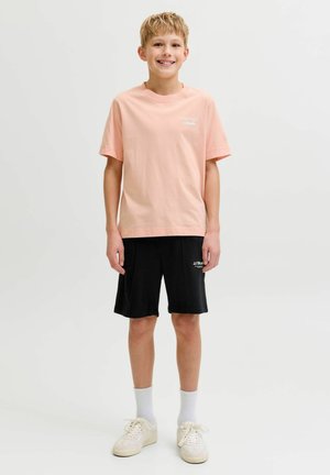 Băiat zâmbitor purtând un tricou roz deschis, pantaloni scurți negri, șosete albe și adidași albi, stând pe un fundal simplu și deschis la culoare.