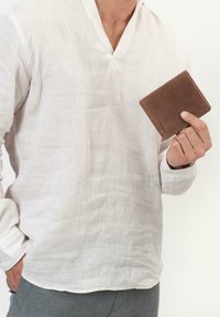 Bruine leren portemonnee vastgehouden in de hand, met een gladde textuur en subtiele stiksels, op de achtergrond van een licht linnen overhemd.