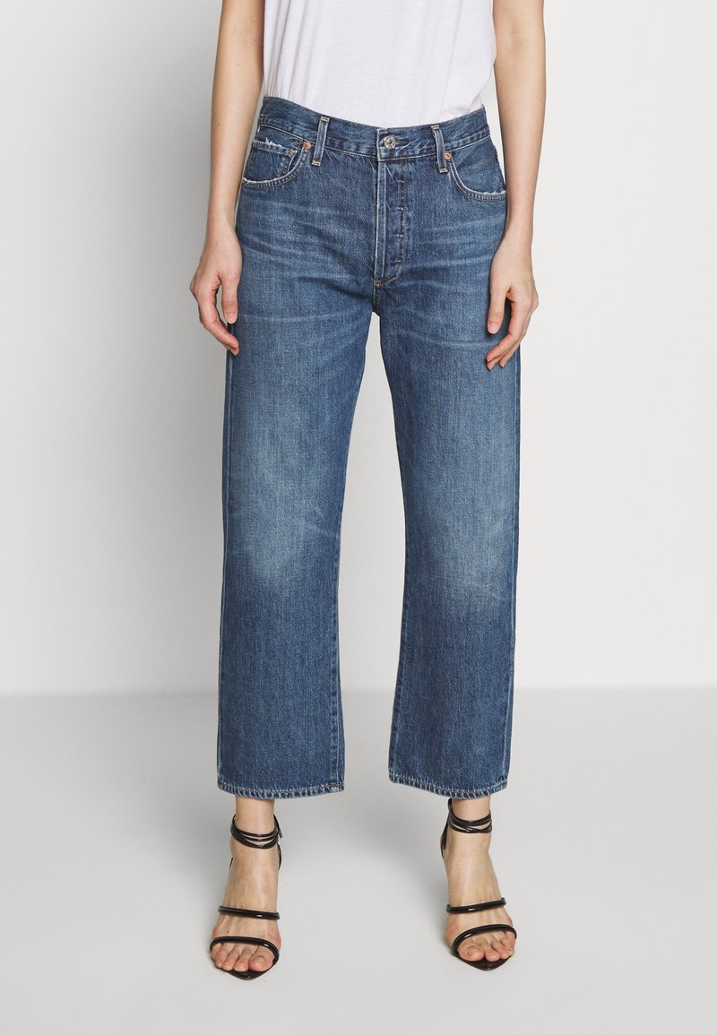 Kvinna i skurna blå jeans med blekta detaljer, matchade med svarta högklackade sandaler med remmar och en vit topp, stående mot vit bakgrund.