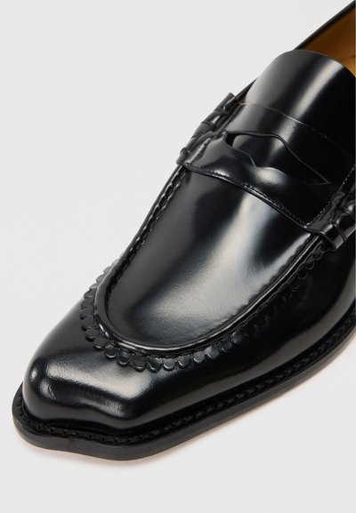 Sapatilha de couro preto com acabamento polido, apresentando um bico arredondado, costura decorativa em viés e uma sola de perfil baixo.