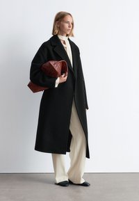 Schwarzer Wollmantel mit taillierter Passform, über einer cremefarbenen Bluse und beigen Hosen getragen. Hält eine strukturierte dunkelrote Handtasche. Schwarze spitze Schuhe.