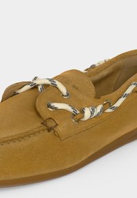 Καφέ καστόρινo boat shoe με λευκά και μαύρα ριγωτά κορδόνια που περνούν από μεταλλικά κρικιά, με ραφές μοκασίνου και μαλακή σόλα.
