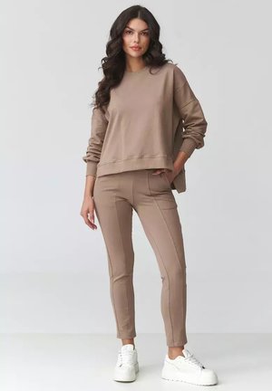 Kobieta o długich ciemnych włosach, ubrana w beżową, oversize'ową bluzę, dopasowane beżowe spodnie i białe sneakersy na platformie, stojąca z jedną ręką w kieszeni.