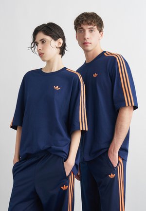 Dve osebi, oblečeni v ujemajoče se Adidas trenirke temno modre barve z oranžnimi črtami, stojita pred enobarvnim belim ozadjem.