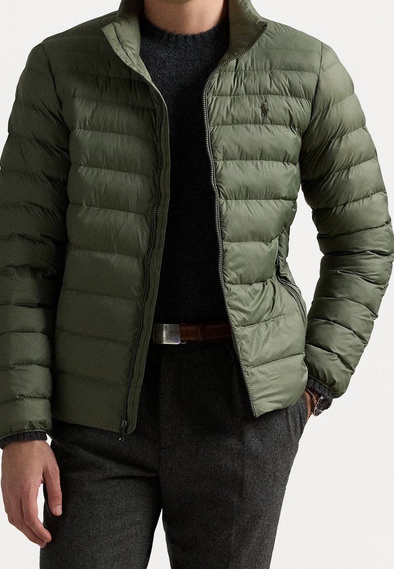 Mężczyzna w rozpiętej oliwkowozielonej pikowanej kurtce, na czarnym swetrze, w szarych spodniach i z brązowym paskiem, z jedną ręką w kieszeni.