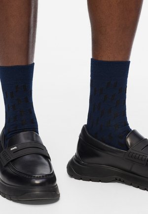 Pies con calcetines de patrón azul marino y mocasines de cuero negro con suelas gruesas sobre un fondo blanco.