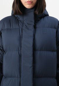 Tamsiai mėlynas pūkinis džemperis su aukštu apykakle, siuvinėtomis detalėmis ir lygiu paviršiumi. Yra minimalistinė logo ant krūtinės ir priekinis užtrauktukas.
