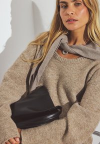 Beige stickad tröja med rund halsringning, grå scarf hängande runt halsen, och en svart läderväska med en kurvad form som hålls i armen.