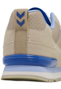 Beige Wildleder-Sneaker mit blauen Akzenten, strukturiertem Rückenteil, blauem Gummifersen und geflochtenem Riemen mit einem blauen Chevron-Muster.