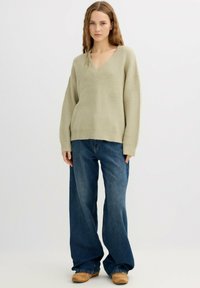 Suéter de punto verde claro con textura y escote en V. Combinado con jeans de corte amplio en azul oscuro y zapatos beige sin cordones. Cabello largo y ondulado.