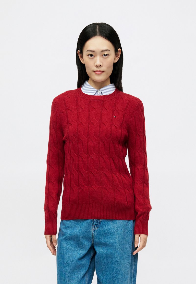 Nuori nainen, jolla on suorat mustat hiukset, pukeutuneena punaisiin palmikkoneuleisiin puseroon raidallisen paidan ja sinisten farkkujen päällä, seisoo valkoista taustaa vasten.