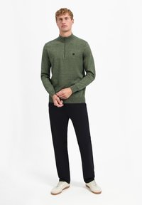 Groene sweater met ritssluiting, gebreid met een textuur, met lange mouwen en een klein logo op de borst, gecombineerd met zwarte broek en witte sneakers.