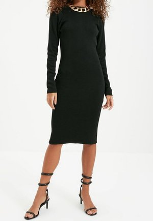 Mujer con un vestido midi negro ceñido de manga larga, un collar grueso de cadena dorada y sandalias negras de tacón con tiras.