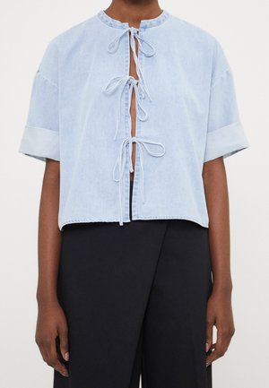 Jasnoniebieska krótka bluzka z denimu z trzema wiązaniami z przodu, połączona z wysokim stanem czarnymi spodniami na ciemnoskórej osobie.