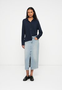 Camisa azul-marinho de botões com mangas compridas, combinada com uma saia de ganga azul-claro com fenda frontal, e sapatos de lona pretos.
